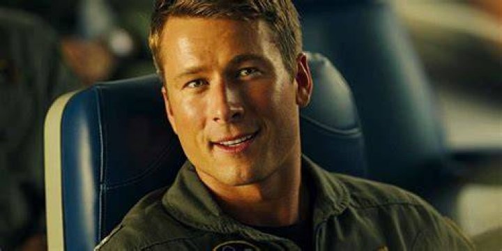 Glen Powell in character as Hangman in Top Gun Maverick wearing a military uniform and holding a pool cue