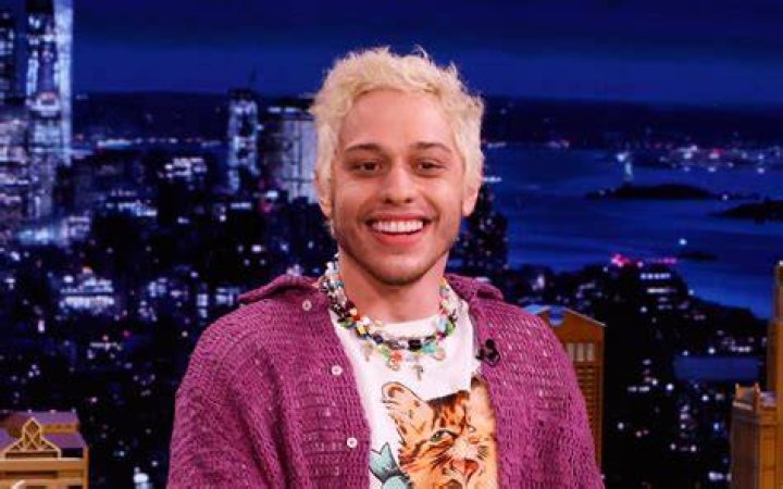 Pete Davidson in a pink shirt and jean jacket against the Weekend Update backdrop on SNL.